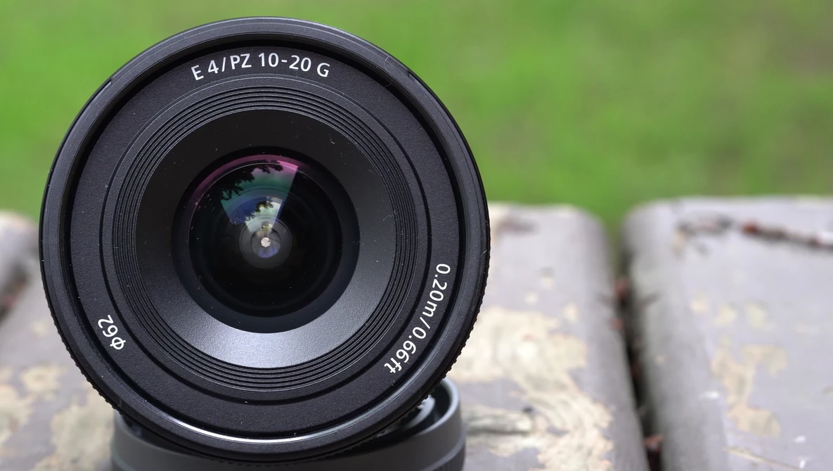 Sony E 10-20mm f/4 PZ G wide-angle zoom lens viewed head-on, mounted on brick surface with blurred green background.