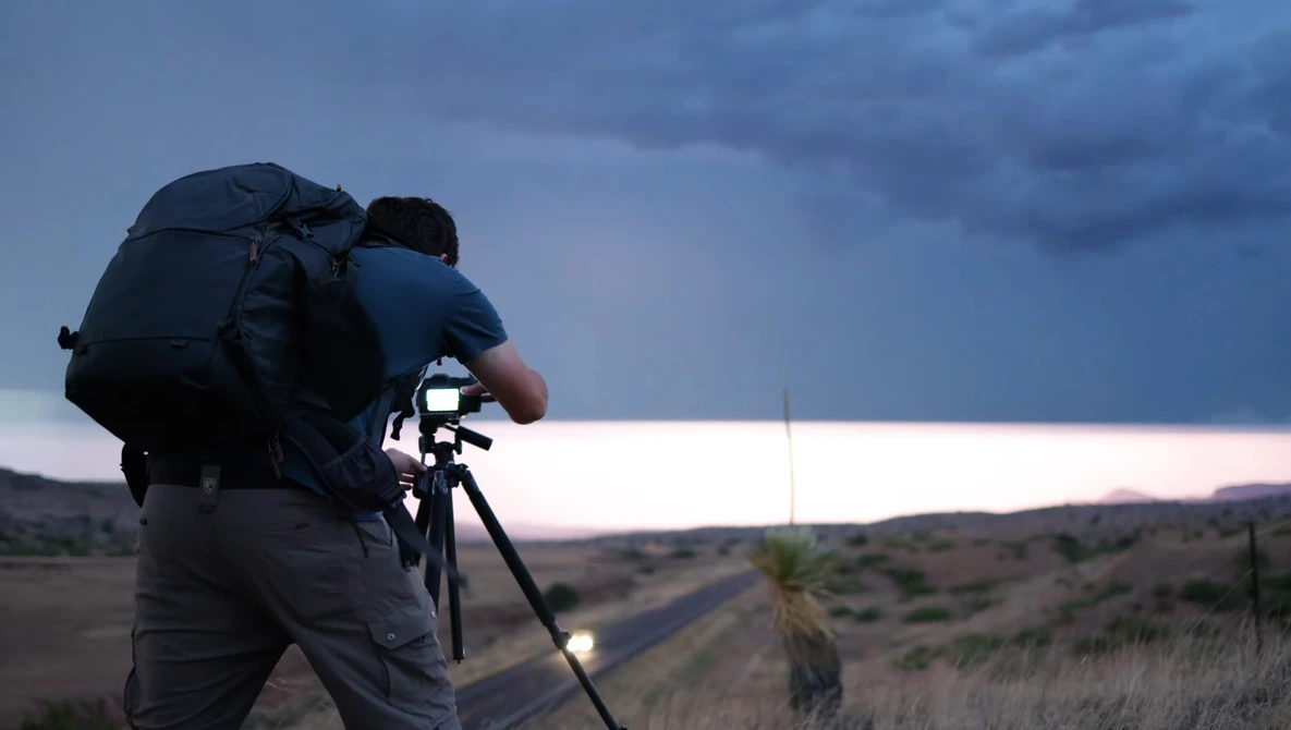 Photographer with large backpack operating a telephoto lens on tripod at dusk overlooking an expansive landscape.