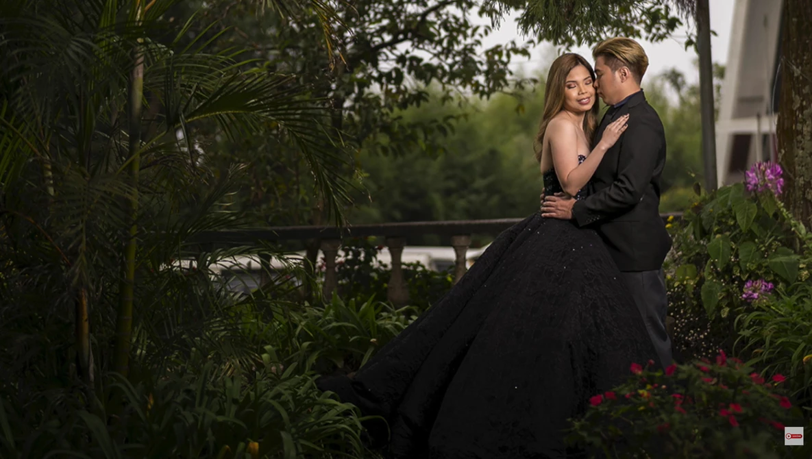 Man in dark suit embracing woman in black ball gown in lush garden setting with flowering plants.