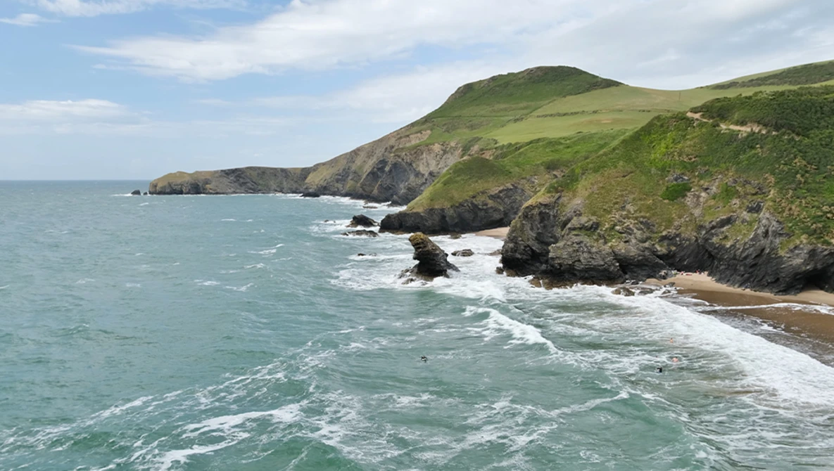 Rugged green coastal cliffs with white-capped waves crashing against rocky outcrops below.