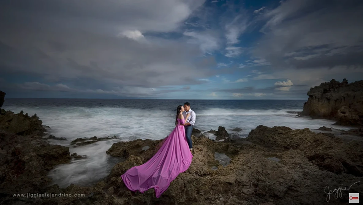 Couple embracing on rocky beach in flowing pink gown with long exposure ocean and dramatic sky.