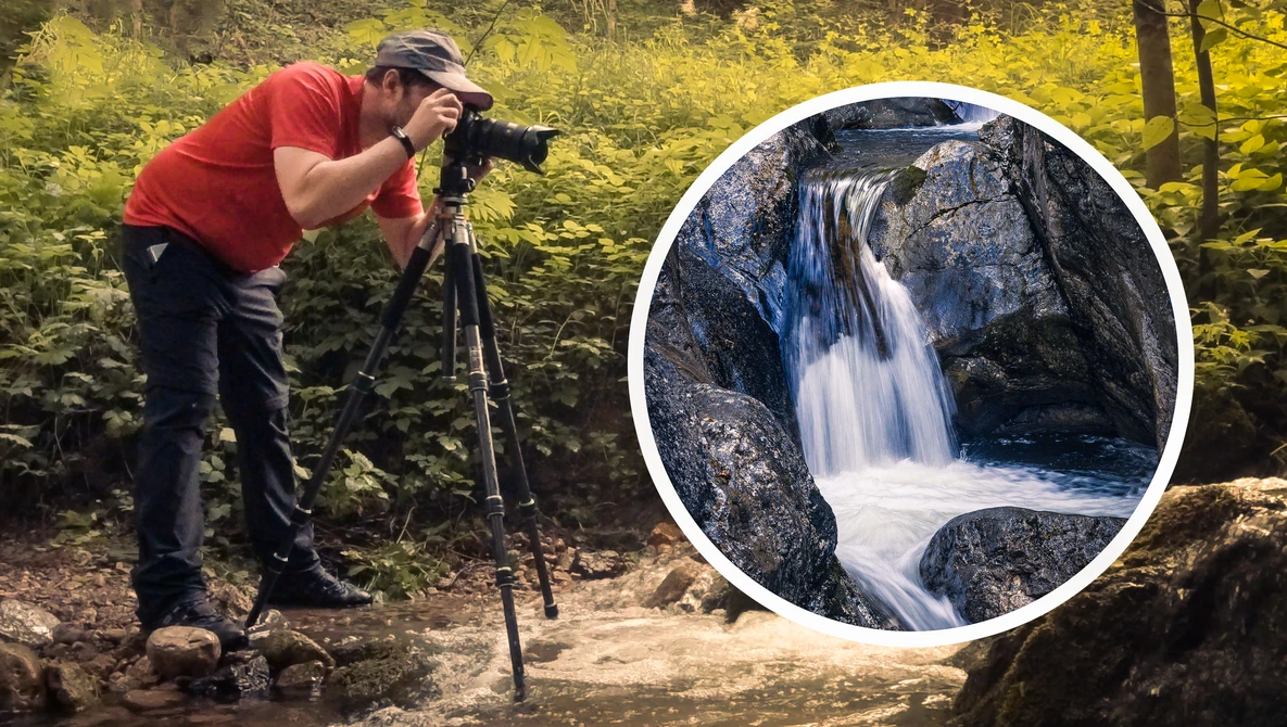 Photographer in red shirt and cap photographing a cascading waterfall in a forested area using a tripod-mounted camera.