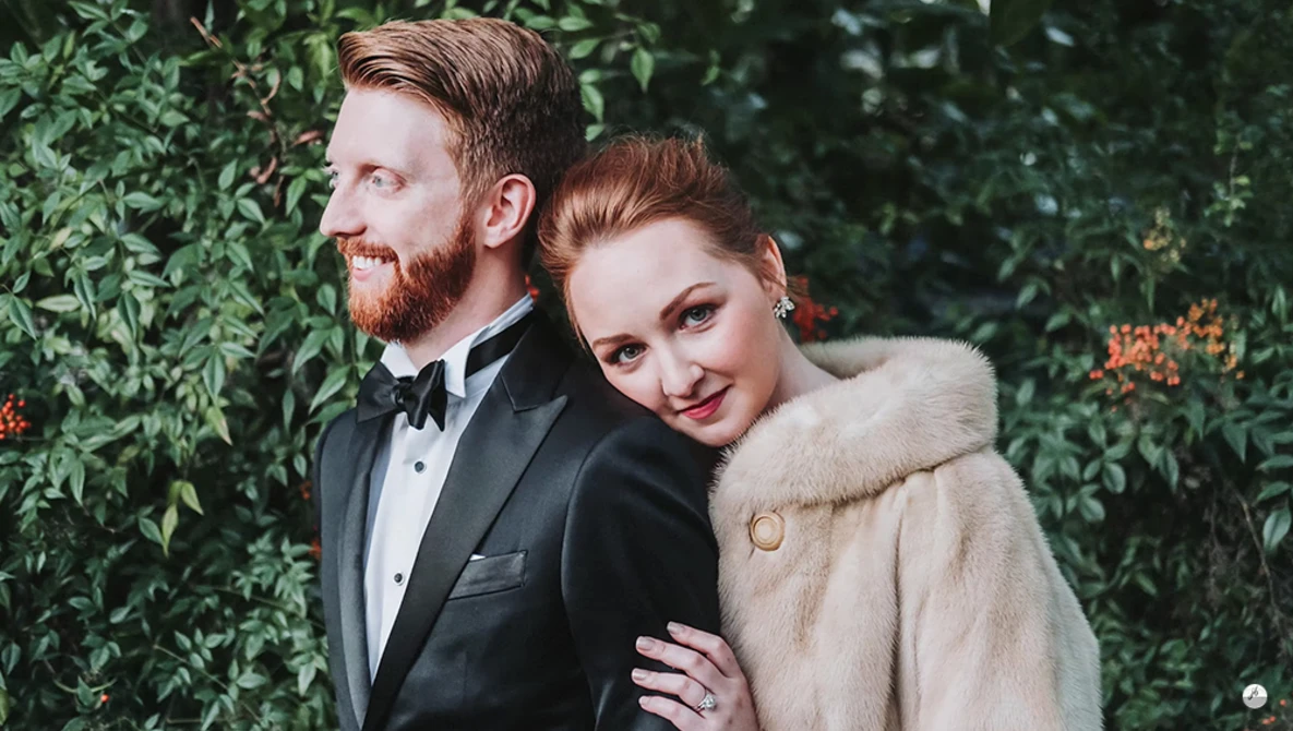 Couple in formal attire posing against a lush green ivy backdrop.