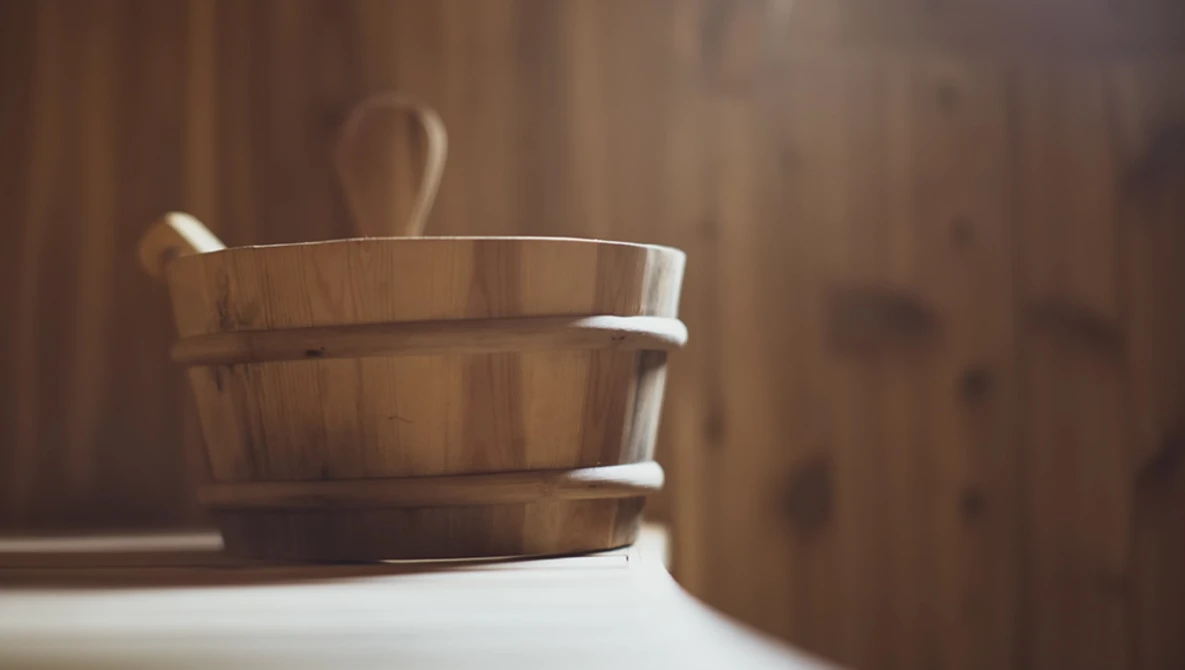 Wooden bucket with metal bands and ladle on white surface against wooden backdrop.