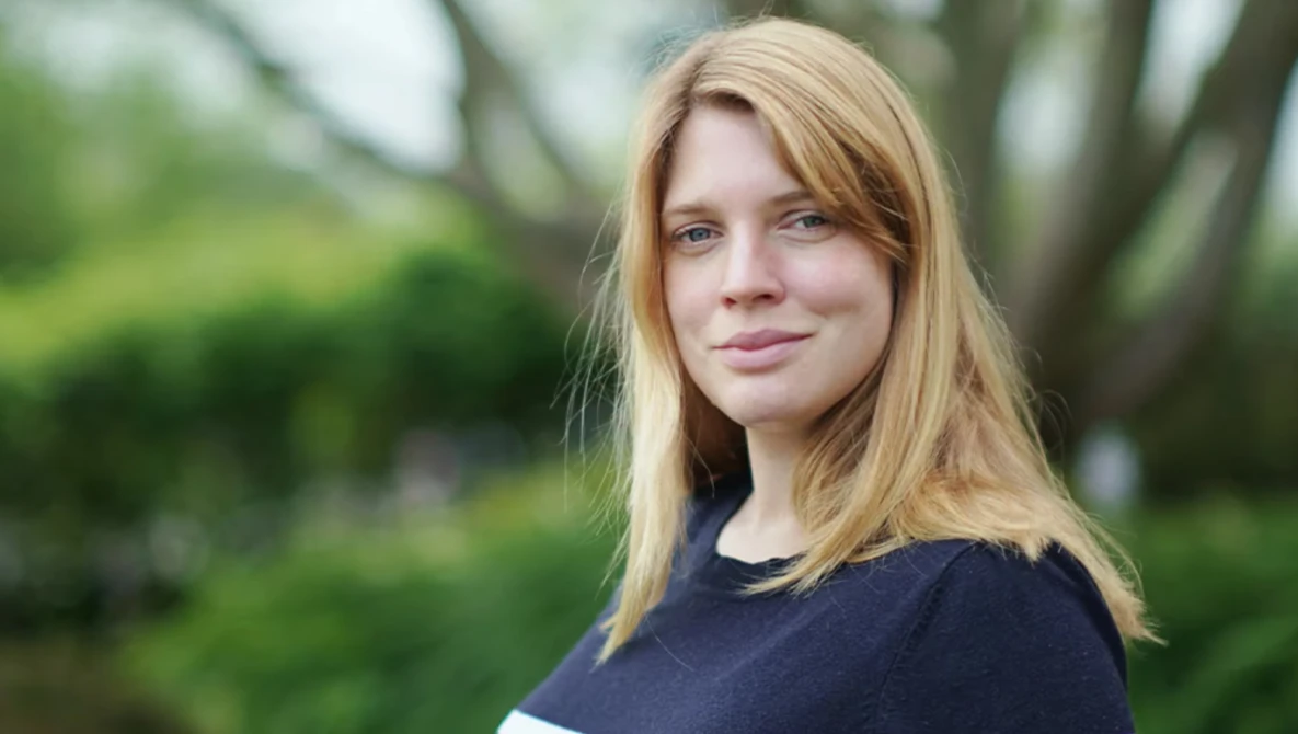 Woman with strawberry blonde hair looking toward camera in outdoor setting with blurred green foliage background.