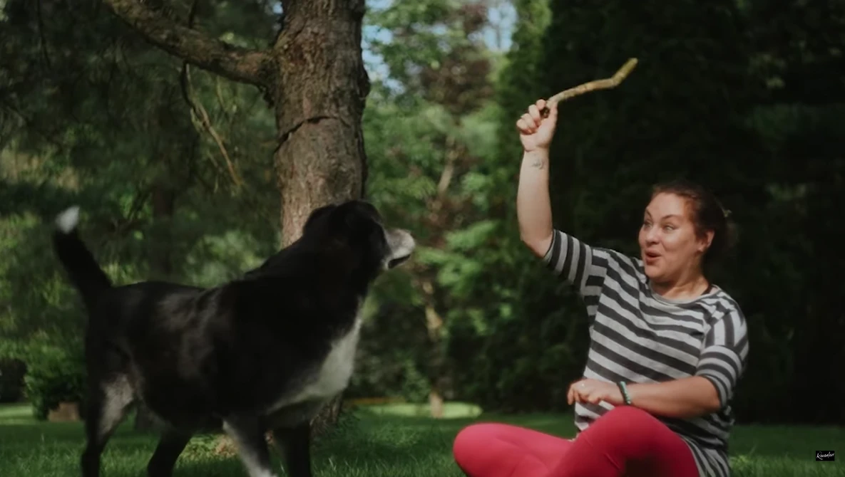 Woman in striped shirt playing fetch with black dog in grassy park setting.