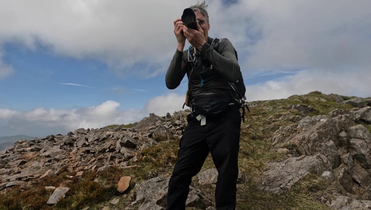 Photographer holding camera up to face while standing on rocky hillside with sparse vegetation.