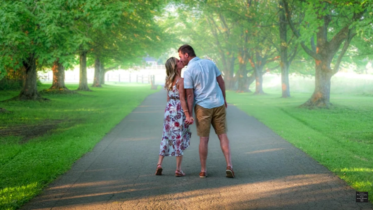 Couple kissing on a tree-lined path with bright sunlight filtering through green foliage.