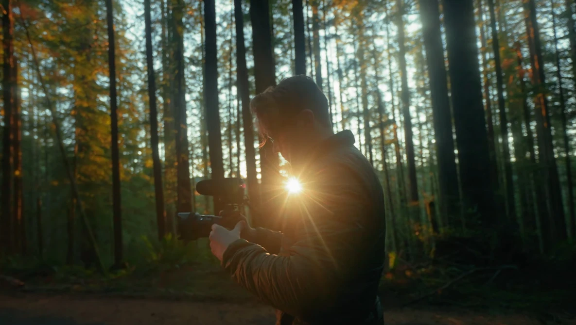 Photographer holding a camera with bright sunlight flaring through the lens in a forest setting.