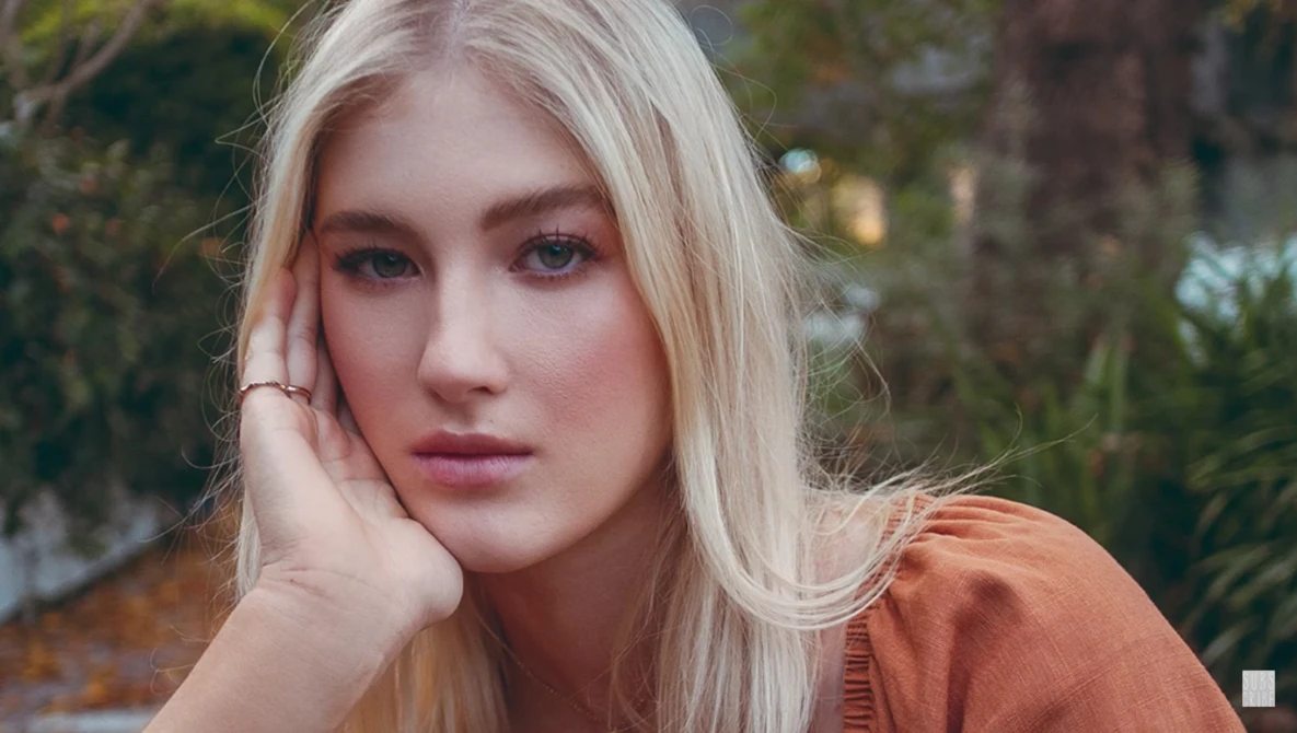 Platinum blonde woman with hand to face gazing at camera, wearing rust-orange cardigan.