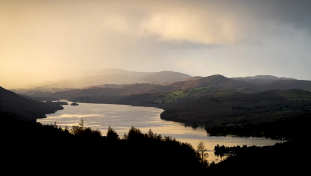 Golden-hour landscape with a still lake surrounded by forested hills and distant mountains.