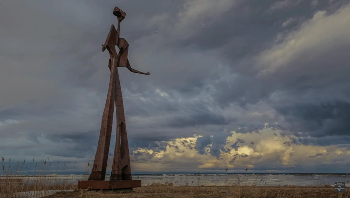 Tall rusted metal sculpture of a figure with outstretched arm against dramatic cloudy sky and water.