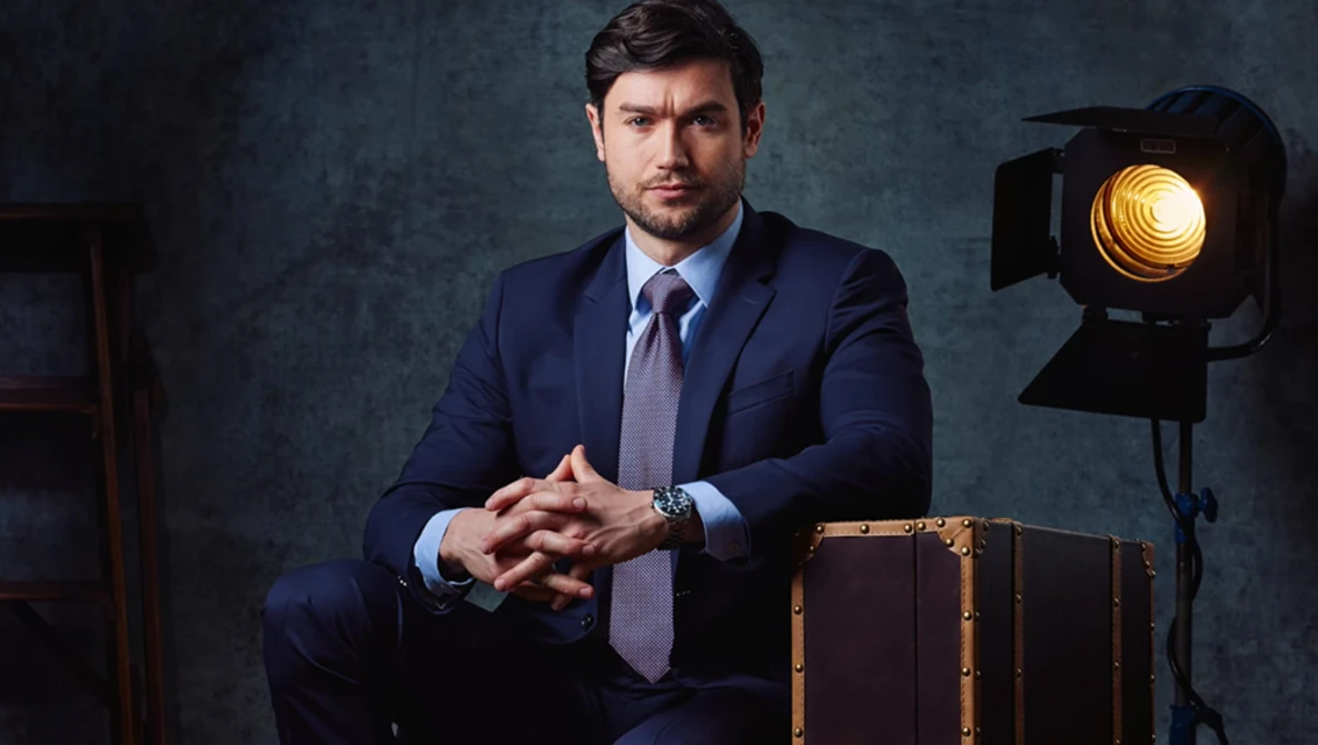 Professional man in navy suit seated next to vintage leather suitcase with studio lighting setup visible.