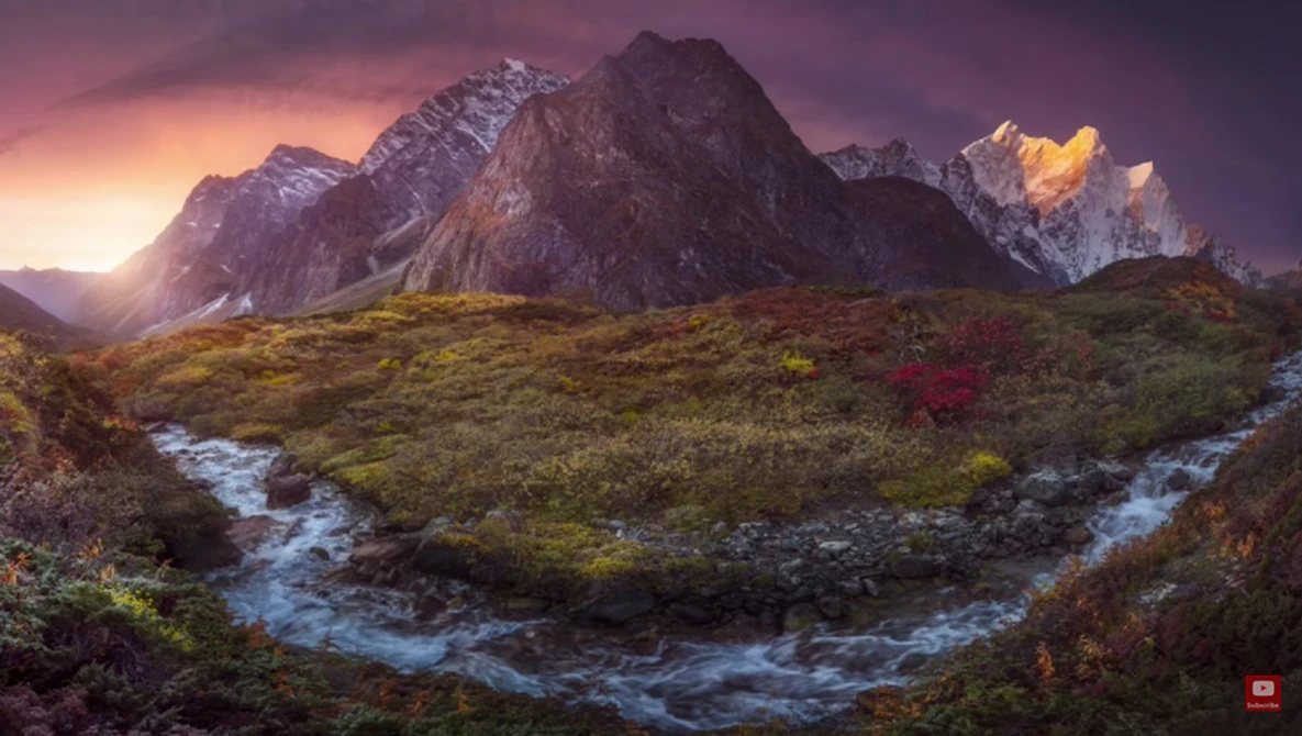 Snow-capped mountain peaks rise above a moss-covered valley with a winding blue stream at sunset.