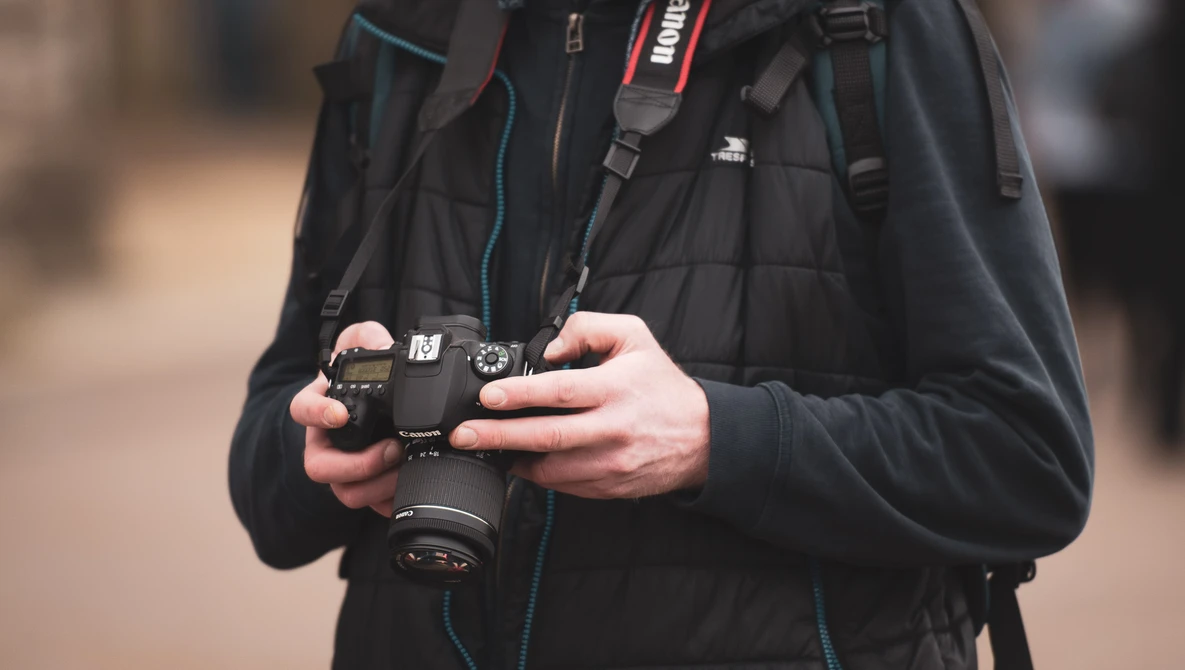 Photographer holding a DSLR camera with a telephoto lens attached, wearing a black jacket with camera straps.