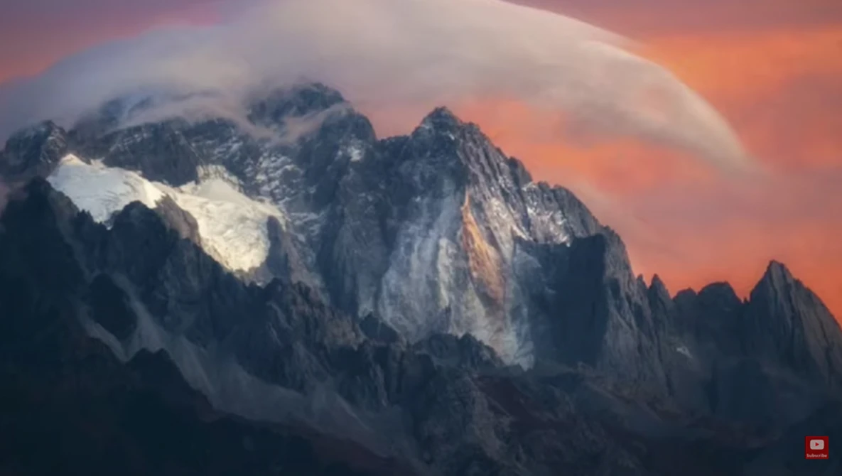 Snow-capped mountain peaks illuminated by dramatic orange and pink sunset light beneath layered clouds.
