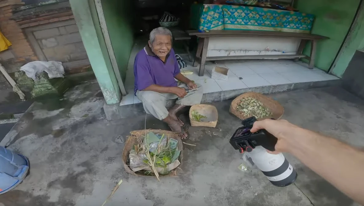 Elderly person in purple shirt preparing fresh vegetables outdoors on concrete ground.