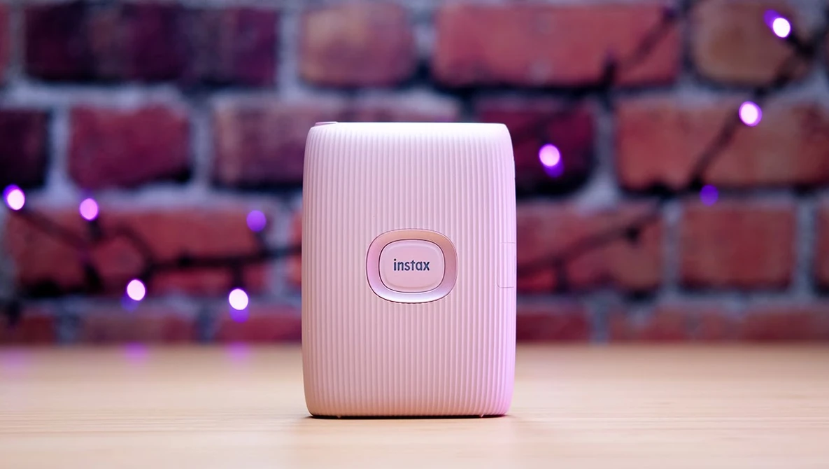 Pink Instax instant camera printer on wooden surface with brick wall and purple lights in background.
