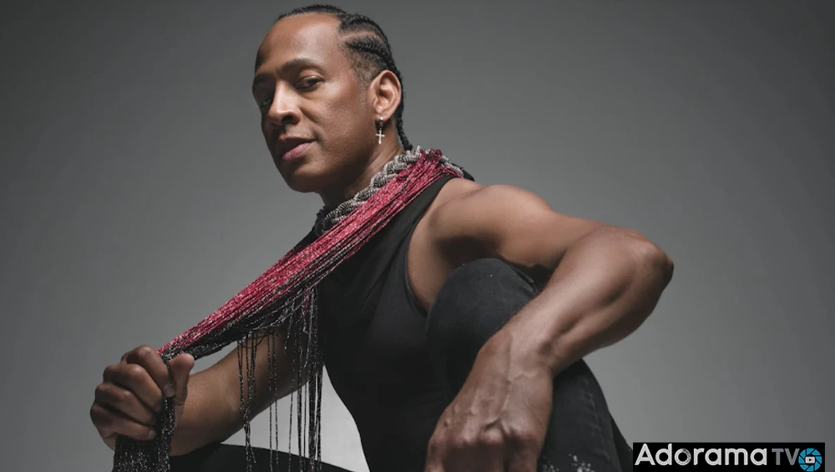 Shirtless man in profile holding a beaded pink and red shoulder scarf against gray background.
