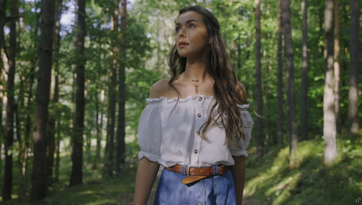 Woman in off-shoulder white blouse and denim skirt standing in a sunlit forest.