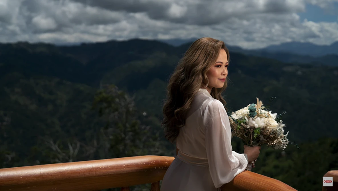 Woman in light blouse holding white flowers at wooden railing overlooking forested mountains.