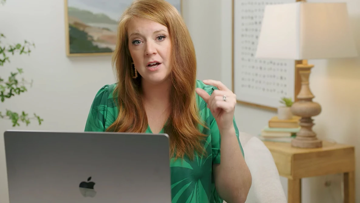 Woman with reddish-blonde hair in green shirt gesturing while speaking at laptop during video call.