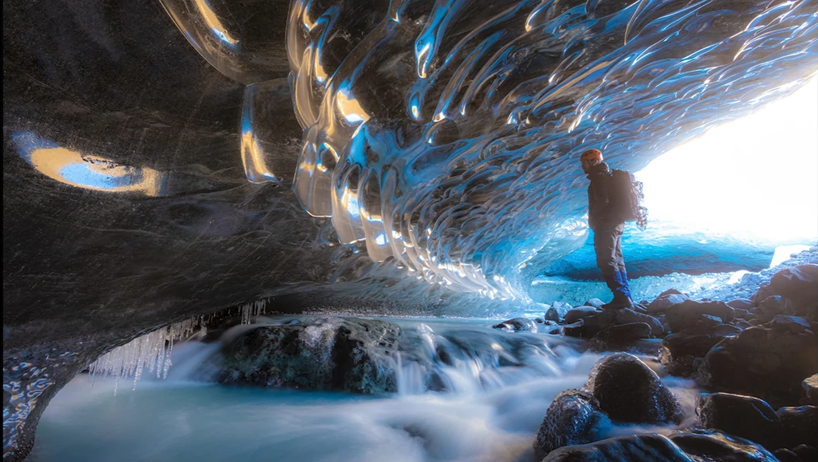 Hiker standing in an ice cave with turquoise glacial stream flowing beneath textured blue ice formations.