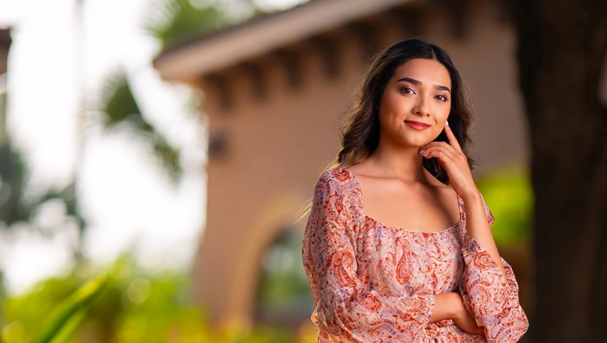 Woman in coral patterned top posing outdoors with blurred residential architecture in background.