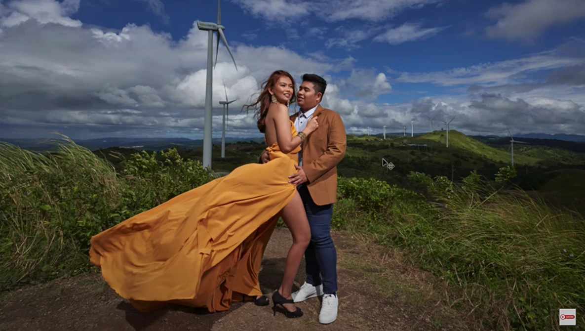 Couple posing together in a rural landscape with a wind turbine and rolling hills in the background.