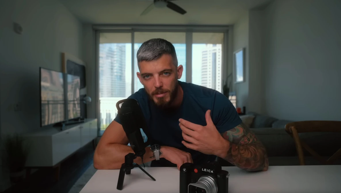 Man with tattoo gesturing while speaking at table with professional camera and microphone setup.