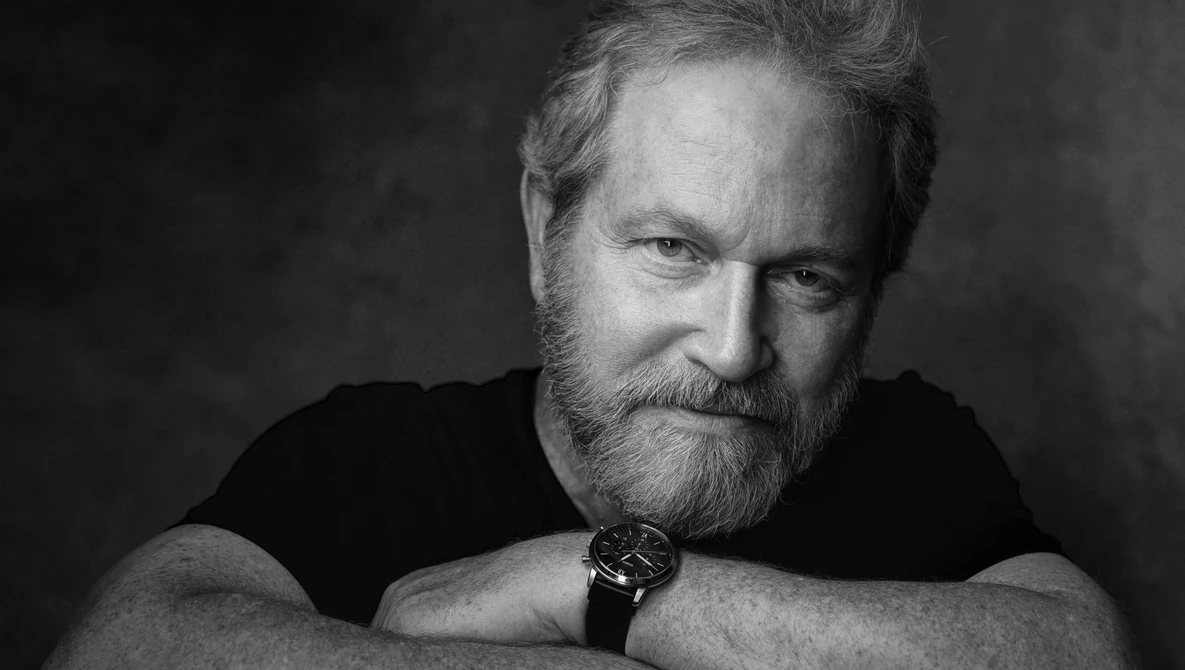 Bearded man in black shirt with watch, posed against dark background in studio portrait.