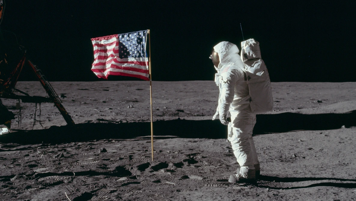Astronaut in white spacesuit standing on lunar surface next to American flag.