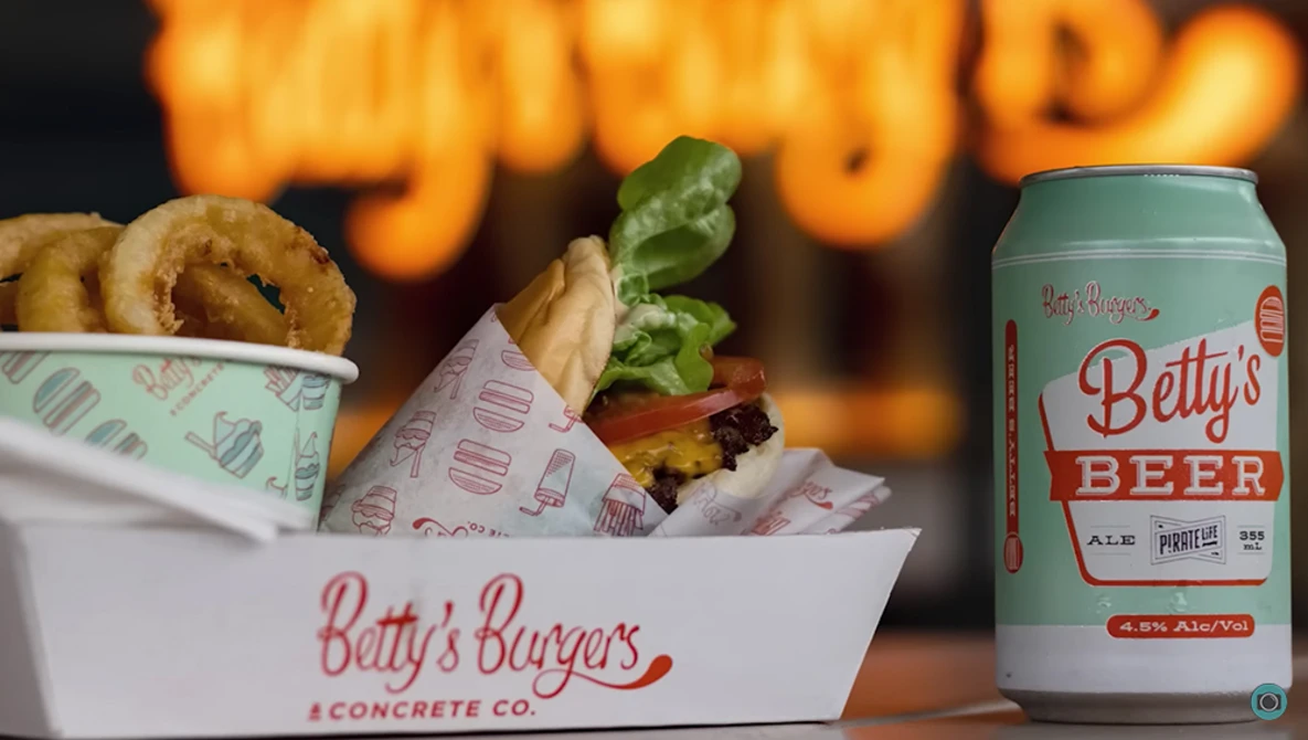 Onion rings and burger in branded paper containers with a beer can on dark background with warm bokeh lighting.
