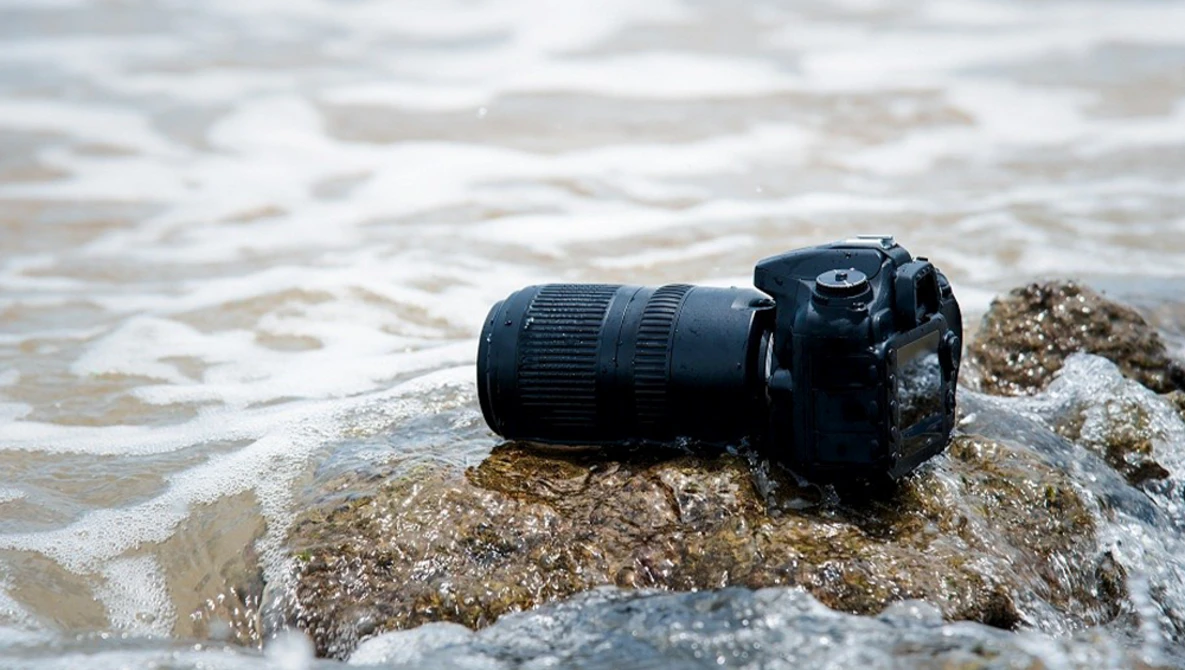 DSLR camera with telephoto lens resting on a rocky beach as waves wash over it.