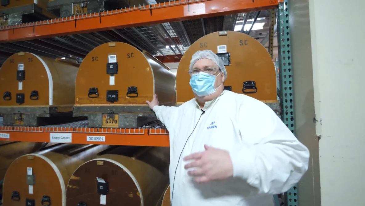 Man in white lab coat and blue mask gestures toward industrial equipment with tan-colored dome-shaped components on orange metal shelving.