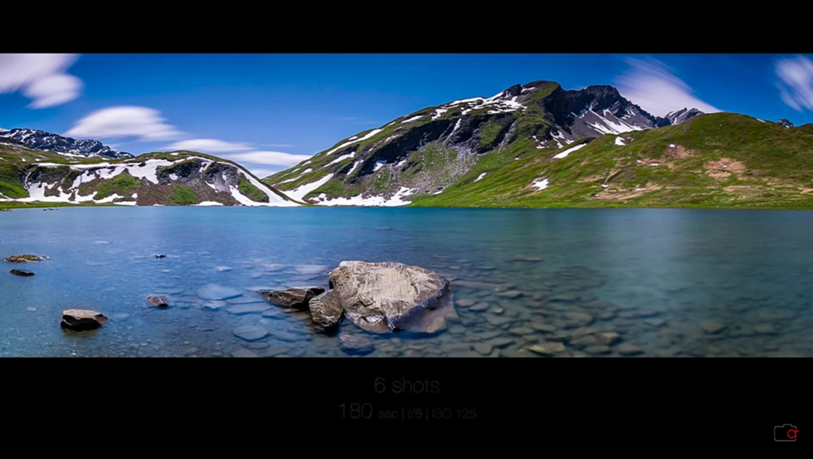 Alpine lake with turquoise water, rocky shores, and snow-capped mountain peaks under blue sky.