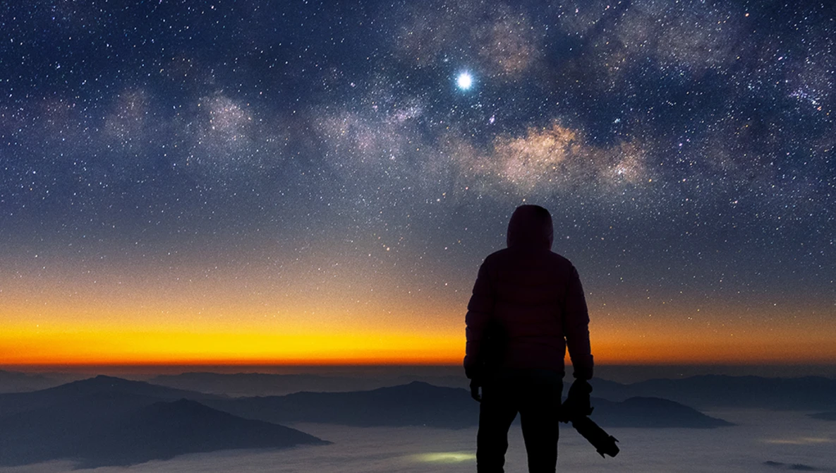 Silhouetted hiker standing on a mountain ridge at twilight, gazing toward a starry sky with the Milky Way visible above an orange sunset horizon.
