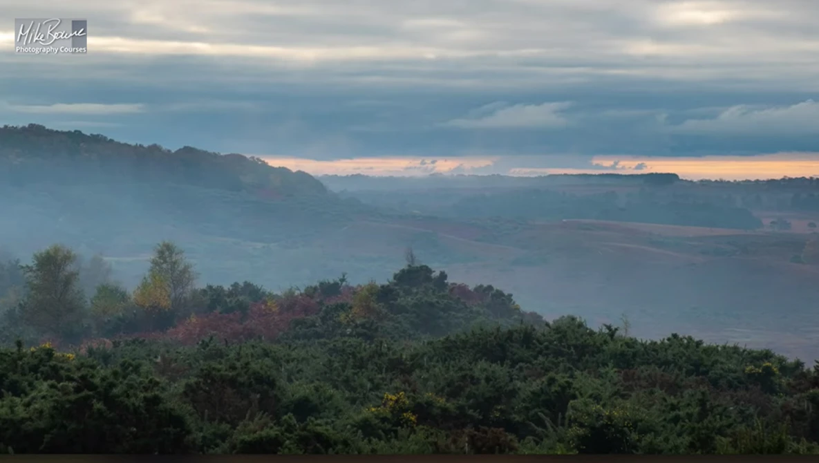 Misty mountain valley at dawn with layered forested ridges and soft golden light breaking through clouds.