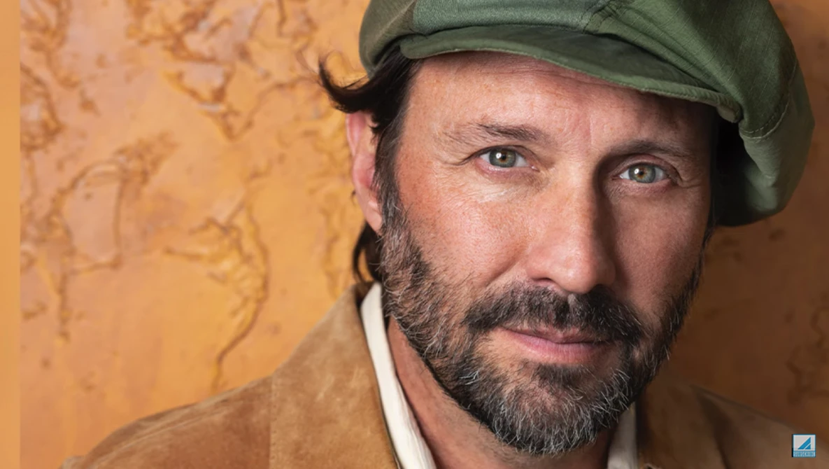 Portrait of a bearded man wearing a green cap against a textured golden-yellow background.
