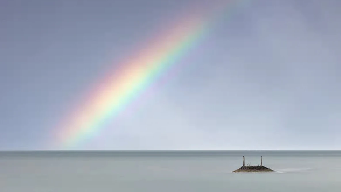 Vivid rainbow arcing over calm, misty water with a small rocky island in the distance.