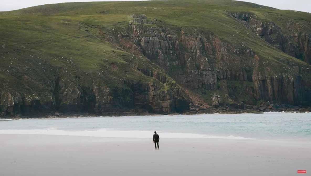 Solitary figure in dark wetsuit standing on pale sand beach beneath towering green hillside with exposed basalt columns.