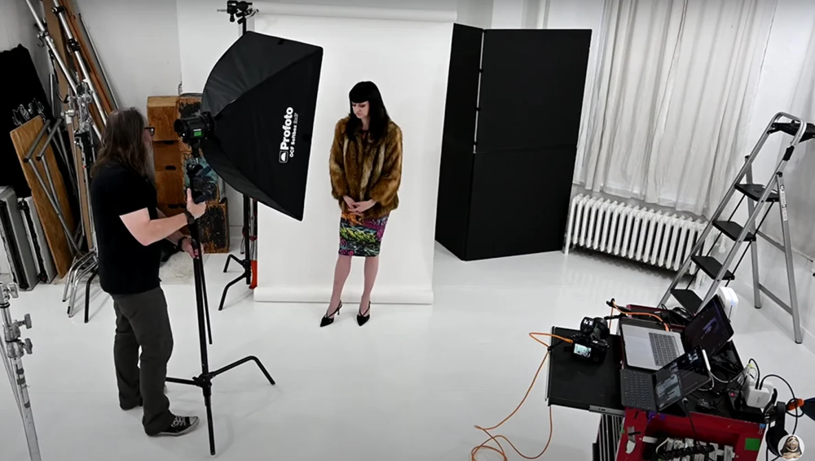 Professional studio setup with photographer operating camera on tripod, subject in brown jacket and patterned skirt positioned under softbox lighting, black reflectors and additional equipment visible.