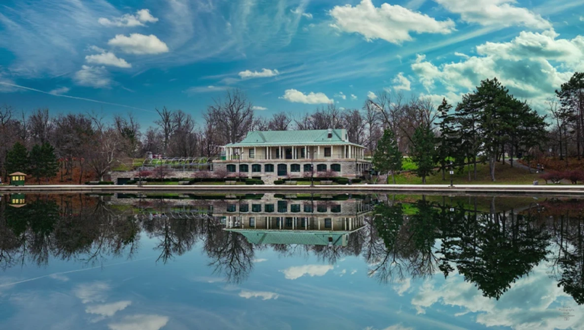 White plantation-style house reflected in still water with bare trees and evergreens along the shoreline.