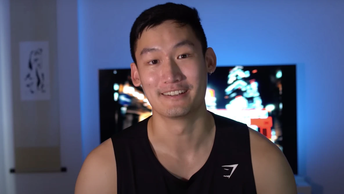 Young man in black sleeveless shirt smiling at camera in blue-lit room with monitors in background.