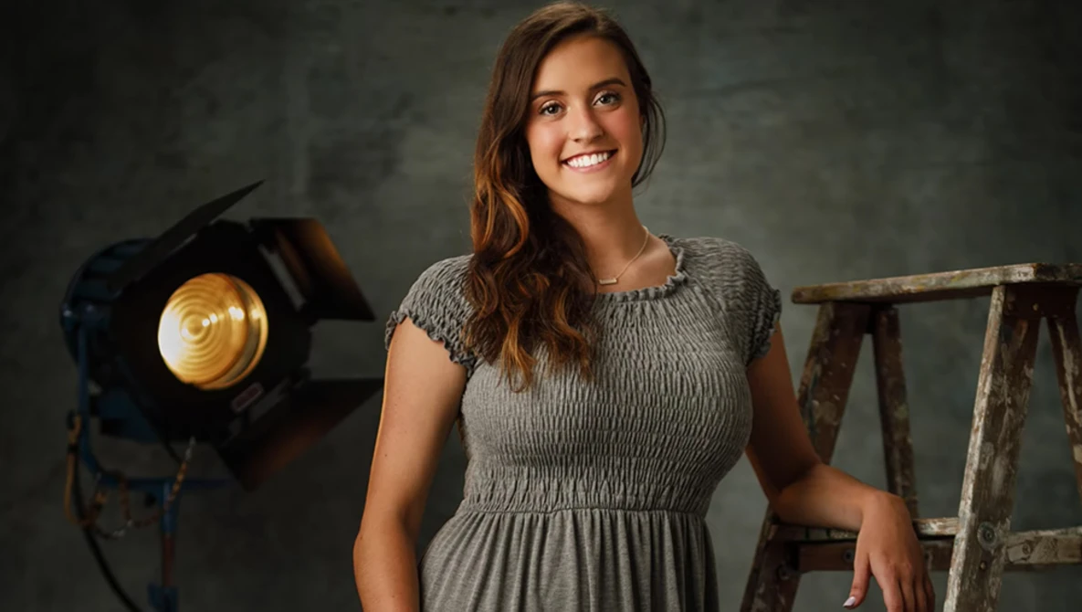 Woman in gray dress smiling at camera during studio portrait session with professional lighting equipment visible.