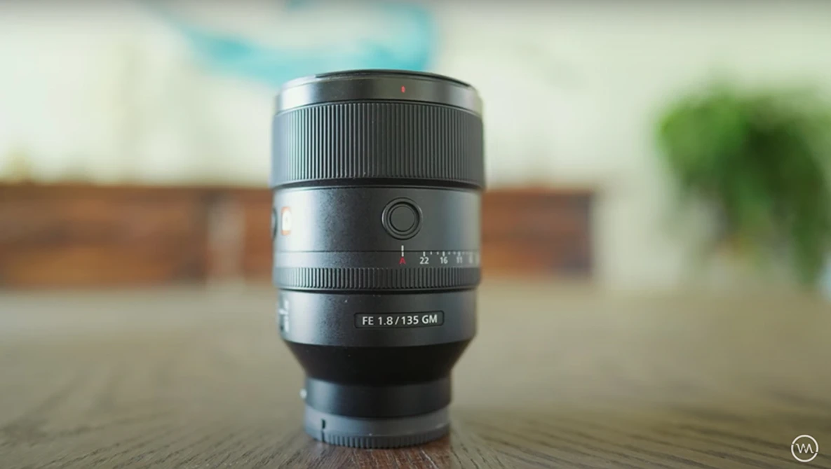 Sony FE 1.8/135 GM lens displayed upright on a wooden surface with blurred interior background.