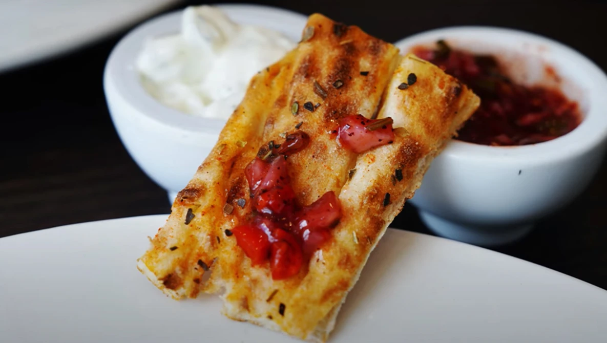 Toasted bread slice topped with melted cheese and red peppers, served with white dipping sauces and a red condiment bowl.