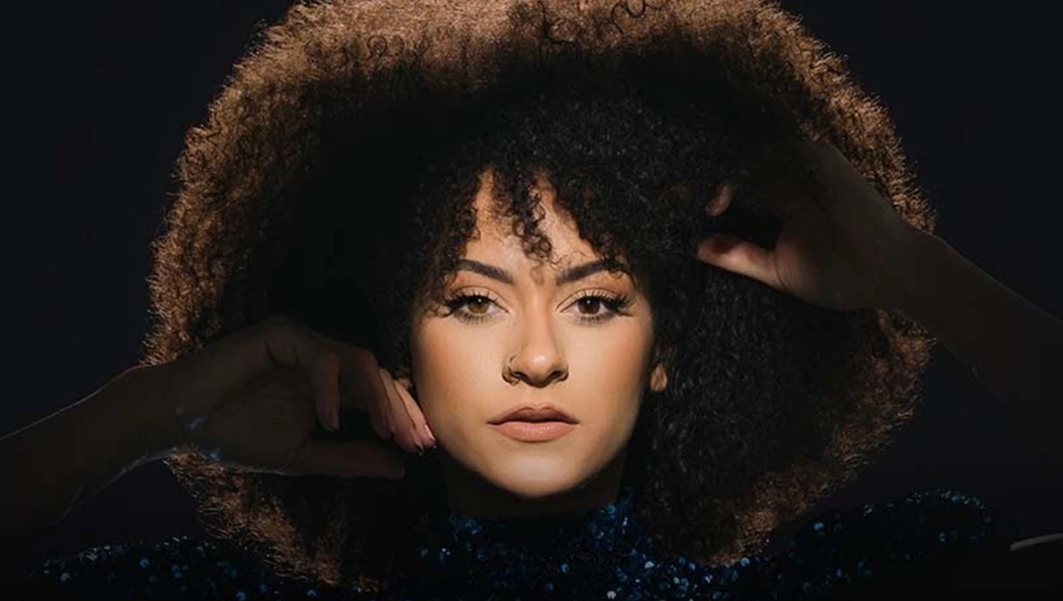 Portrait of a woman with textured dark curly hair and bangs, hand raised to her head, against a dark background.