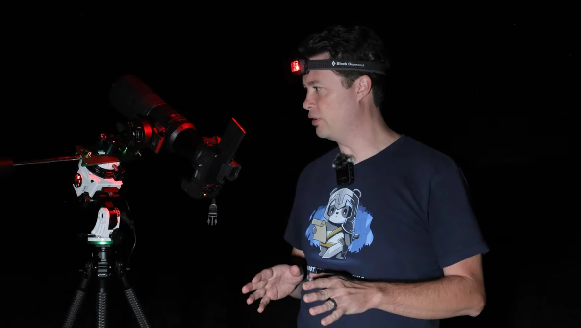 Photographer wearing headlamp demonstrates camera setup with red-lit telescope equipment in dark studio.