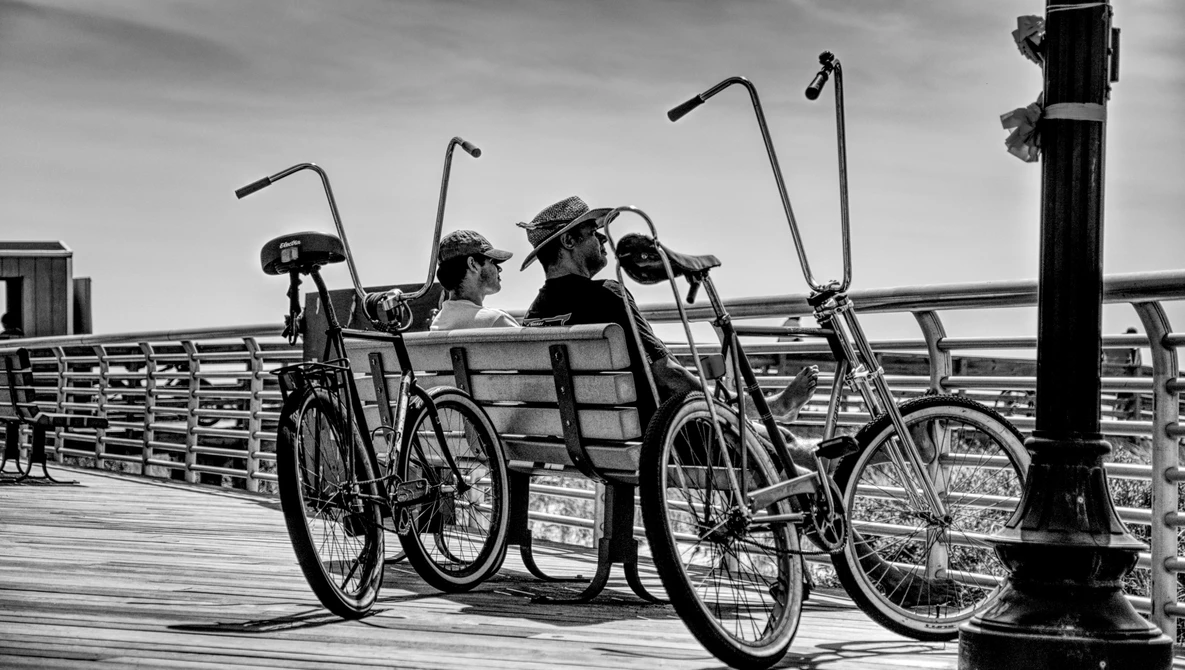 Two people sitting on a bench between bicycles on a wooden pier, photographed in black and white.